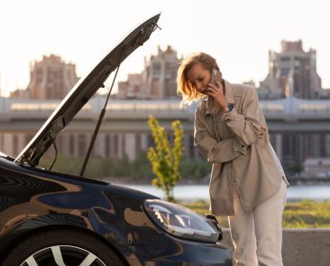 Helpless woman driver calling for help assistance looking at broken down car with opened hood, talking on mobile phone, stopped at the roadside. Doesn't understand what happened.